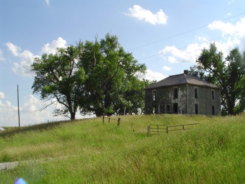 Abandoned house on a grassy hill, surrounded by trees and under a blue sky with fluffy clouds.