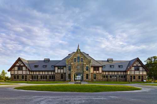 A large stone building with a peaked roof, surrounded by grass and a circular driveway under a cloudy sky.