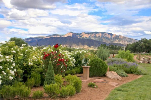 A scenic garden with colorful flowers and shrubs, set against a backdrop of mountains and a cloudy sky.