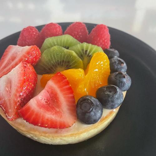 A fruit tart topped with strawberries, raspberries, kiwi, blueberries, and orange slices on a black plate.