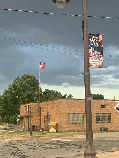 A brick building with a flag and a street banner under a cloudy sky, surrounded by greenery.