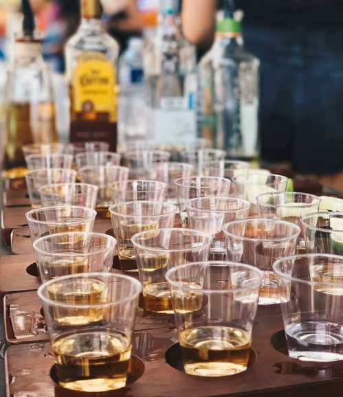 A table with multiple clear plastic cups filled with golden liquid, surrounded by bottles and lime wedges.