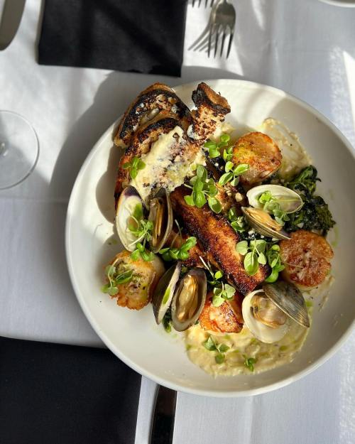 A beautifully plated dish featuring grilled fish, clams, scallops, greens, and creamy sauce, garnished with microgreens.