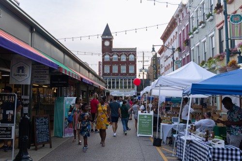 A bustling market street with colorful tents, people shopping, and a historic building in the background under string lights.