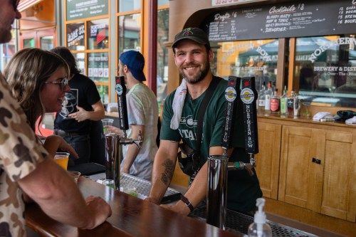 A smiling bartender serves drinks at a lively brewery, with patrons enjoying the atmosphere in the background.