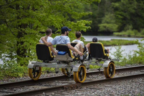 Four people ride a rail bike along a scenic route by a river, surrounded by lush greenery.