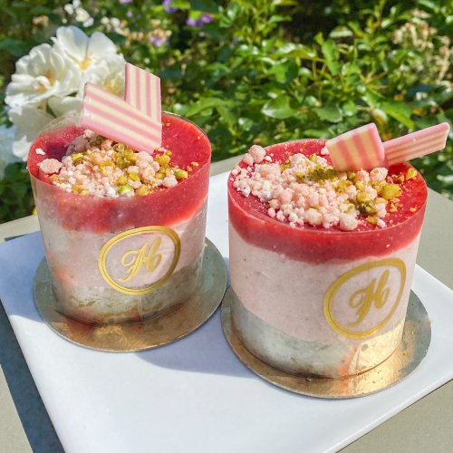 Two elegant dessert cups with layers of red and pink mousse, topped with crushed pistachios and decorative stripes.