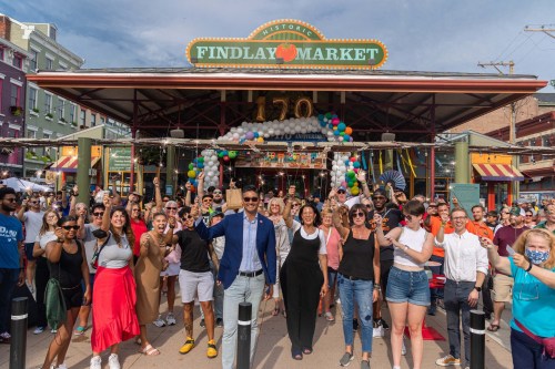 A large crowd gathers outside Findlay Market, celebrating with balloons and smiles on a sunny day.