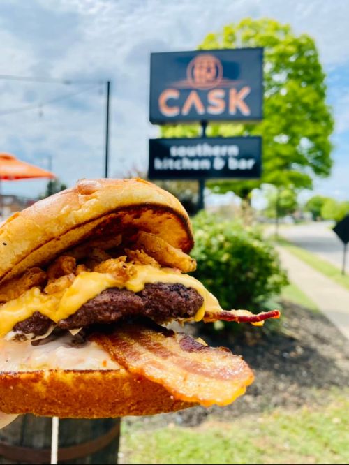 A close-up of a loaded burger with bacon and cheese, with a restaurant sign in the background on a sunny day.