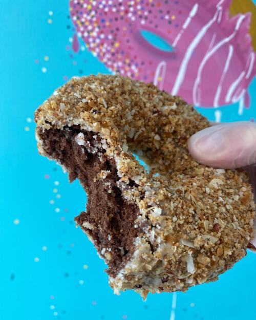 A hand holds a partially eaten chocolate donut coated in crushed nuts, against a colorful, playful background.
