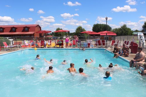 A lively pool scene with children swimming and splashing, surrounded by lounge chairs and umbrellas under a blue sky.
