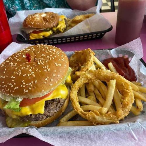 A sesame seed bun burger with cheese, lettuce, and tomato, served with fries, onion rings, and ketchup on a pink table.