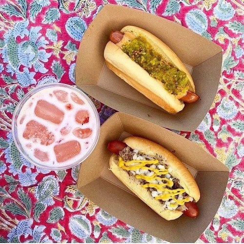 Two hot dogs in paper trays, one with green relish and mustard, the other with chopped onions and yellow mustard, beside a pink drink.