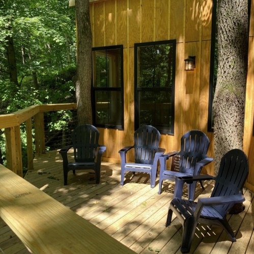 A wooden deck with four blue chairs surrounded by trees and a cabin with large windows.