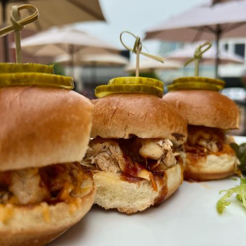 Three mini sandwiches with pulled chicken and sauce, garnished with pickles, served on a white plate.