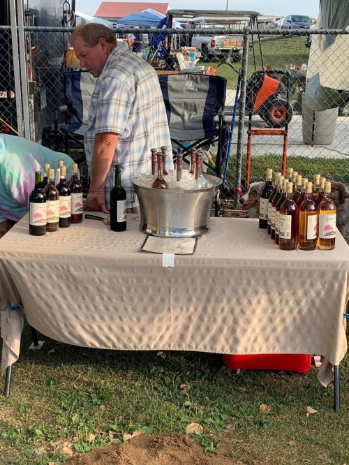 A vendor stands behind a table with bottles of wine and a bucket of ice at an outdoor event.