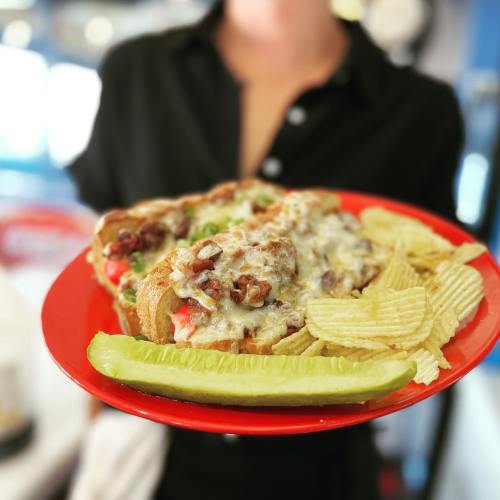 A person holds a red plate with a cheesy sandwich, potato chips, and a pickle, set against a blurred background.
