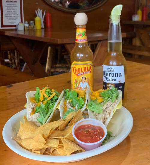 A plate of three tacos with chicken, lettuce, and cheese, served with chips, salsa, and a bottle of Corona beer.