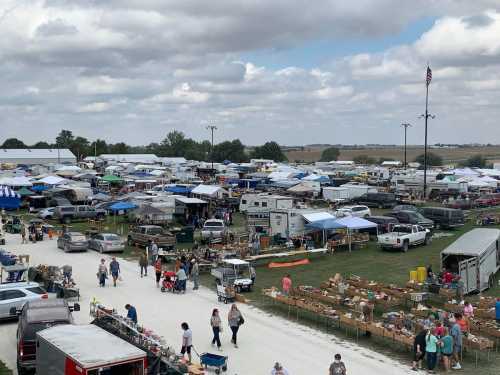 A bustling outdoor market with tents, vendors, and crowds, set against a cloudy sky and open fields.