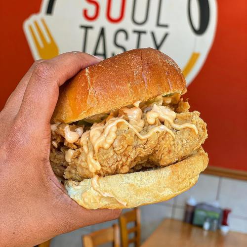 A hand holding a large fried chicken sandwich with sauce, set against a colorful restaurant background.