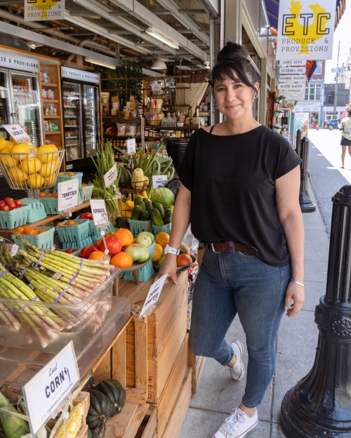 A woman stands by a colorful produce display outside a market, smiling and wearing casual attire.