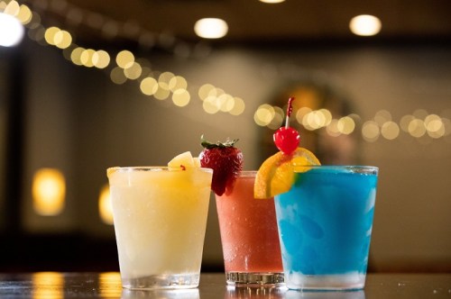 Three colorful cocktails on a bar: a yellow drink with a pineapple, a pink drink with a strawberry, and a blue drink with an orange slice and cherry.