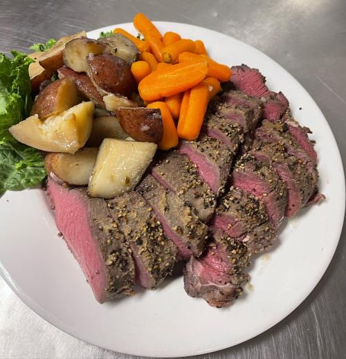 Sliced roast beef with a pepper crust, served with roasted potatoes, carrots, and lettuce on a white plate.