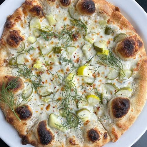 A round pizza topped with cheese, pickles, and fresh dill on a golden crust.
