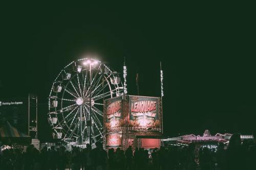 A nighttime carnival scene featuring a large Ferris wheel and colorful game booths illuminated by bright lights.