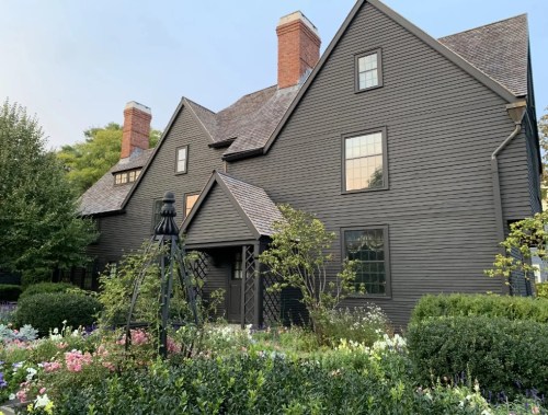 A large, dark-colored house with multiple chimneys, surrounded by colorful flowers and greenery.