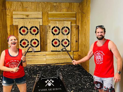 Two people in matching red tank tops stand in an axe-throwing venue, smiling and holding axes in front of target boards.