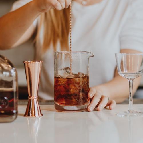 A person stirs a cocktail in a glass beaker, with a jigger and a cocktail glass nearby.