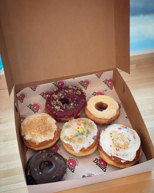 A box of six assorted donuts with various toppings, including sprinkles, frosting, and fruit, on a wooden surface.