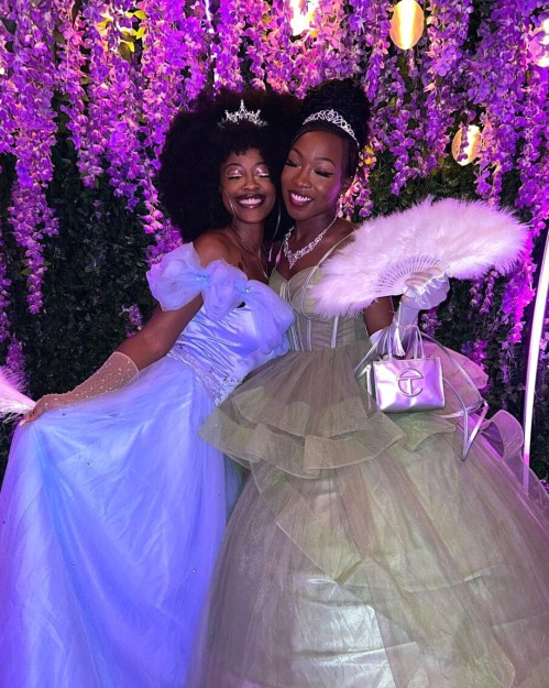 Two women in elegant gowns, one in blue and the other in green, pose joyfully against a backdrop of purple flowers.
