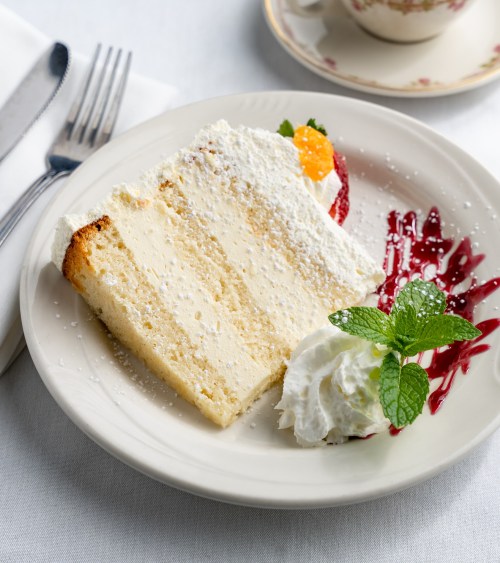 A slice of creamy cake topped with powdered sugar, served with whipped cream, fruit, and a berry sauce on a plate.