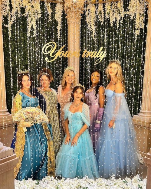 A group of six women in elegant dresses poses together in front of a decorative backdrop with hanging beads and flowers.