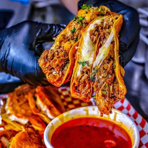 A hand in a black glove holds three loaded tacos above a bowl of red sauce, with more tacos in the background.