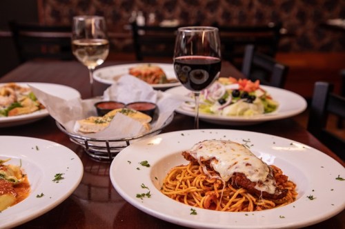 A plate of spaghetti topped with chicken and cheese, surrounded by salads, bread, and glasses of red and white wine.