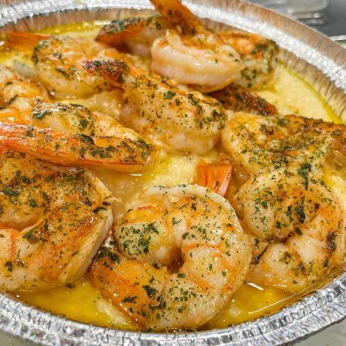A close-up of shrimp cooked in a buttery sauce, served over creamy grits in a silver foil container.