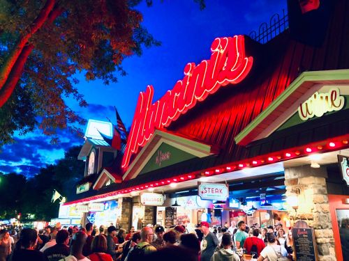 A lively outdoor scene at night with a bustling crowd in front of a restaurant named "Mancini's," illuminated by neon lights.