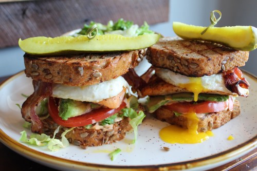 A hearty sandwich with bacon, egg, avocado, and tomato on whole grain bread, garnished with pickles and lettuce.