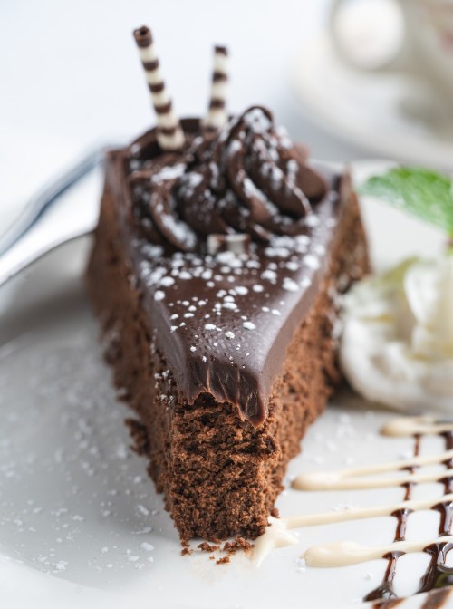 A slice of rich chocolate cake topped with frosting, chocolate curls, and decorative drizzles on a white plate.