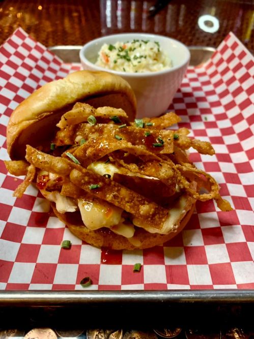 A sandwich with crispy fried onions, melted cheese, and sauce, served with a side of coleslaw in a white bowl.