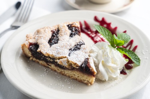 A slice of dessert with a flaky crust, filled with dark fruit, dusted with powdered sugar, and served with whipped cream.