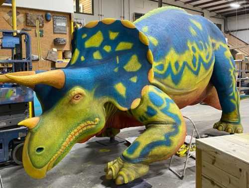 A large, colorful triceratops sculpture with blue, green, and orange patterns, displayed in a workshop setting.