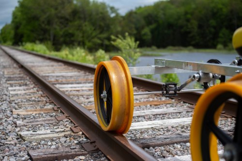 A close-up of a yellow wheel on a rail track, with greenery and a body of water in the background.