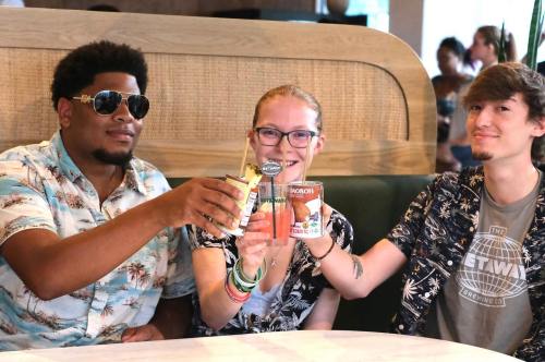 Three friends clink their drinks together at a table, smiling and enjoying a lively atmosphere.