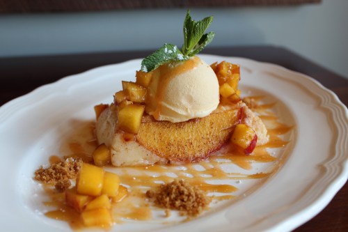 A dessert featuring a slice of cake topped with peach slices, ice cream, and a mint garnish, drizzled with caramel.
