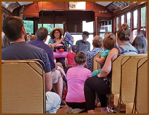 A group of people seated on a vintage train, listening to two presenters at the front.