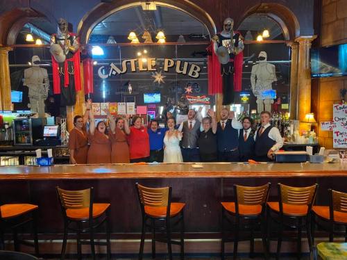 A group of people in formal and casual attire cheer together behind a bar at Castle Pub, celebrating an event.